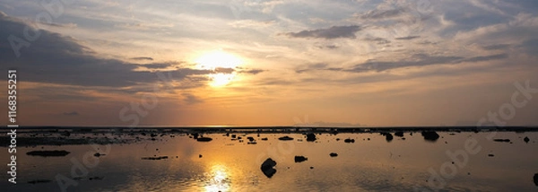 Obraz A serene sunset over a calm, reflective sea with scattered rocks and ripples on the water.