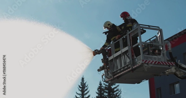 Fototapeta Firefighters stand in a special lifting cradle and put out a fire with a fire hose. The footage shows hard work in extreme conditions. Ideal for themes about rescue operations.