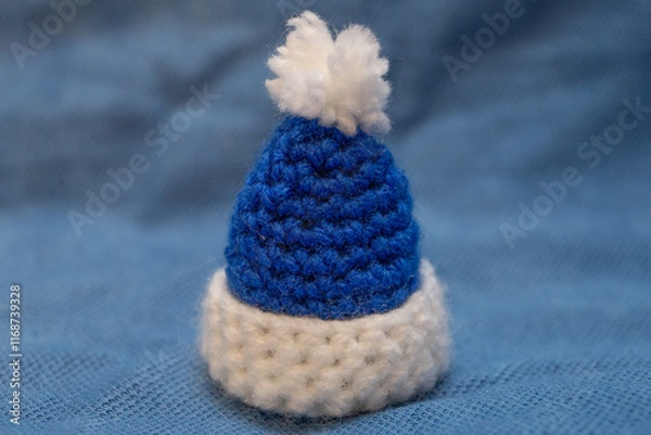 Fototapeta A blue cap with a white pompom for a soft toy, hand-knitted, stands on a blue background.