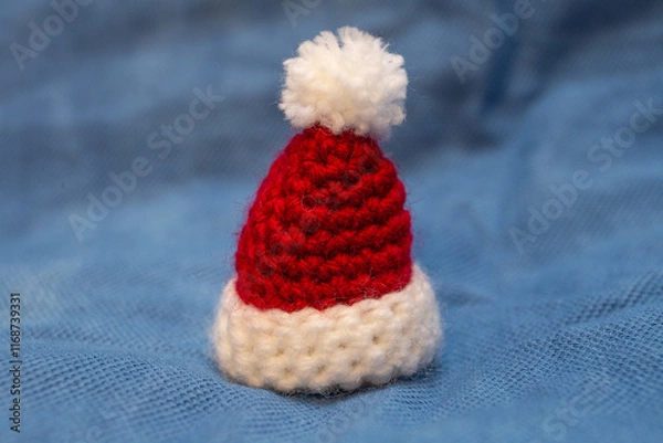 Fototapeta A red cap with a white pompom for a soft toy, hand-knitted, stands on a blue background.