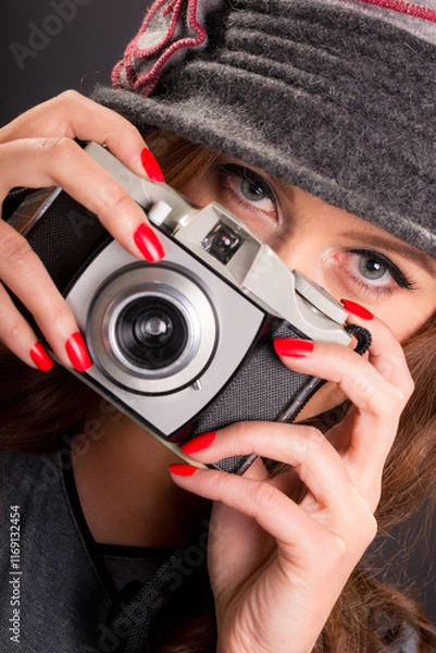 Fototapeta Vintage retro style portrait of a beautiful young woman holding an old film camera, watching and focusing on the viewer ready to take a photograph.