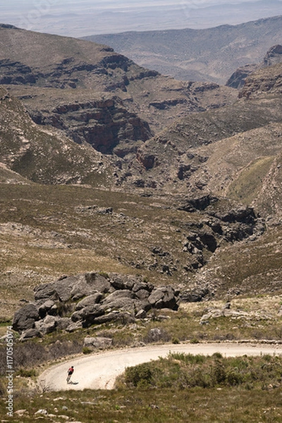 Fototapeta A cyclist descending a steep gravel road down a mountain pass leading to a deep valley.