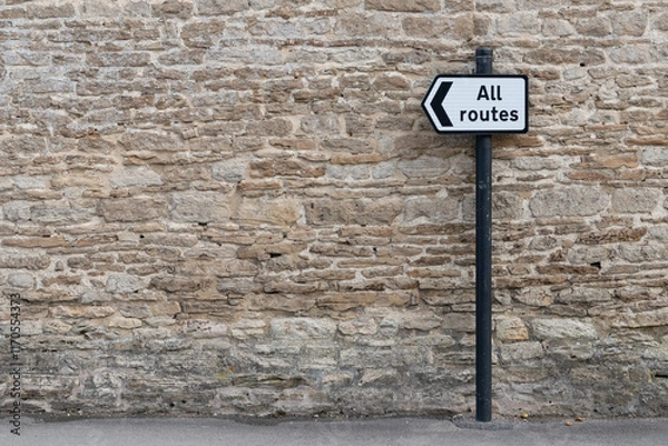 Fototapeta All Routes Road Sign pointing left in front of an old stone wall