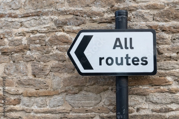 Fototapeta All Routes Road Sign pointing left in front of an old stone wall
