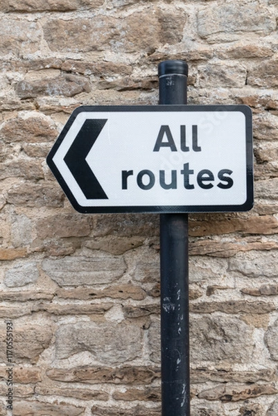 Fototapeta All Routes Road Sign pointing left in front of an old stone wall