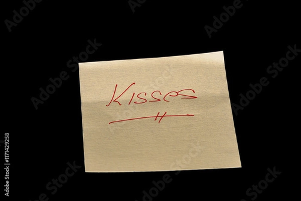 Fototapeta Yellow adhesive paper, similar to a Post-it note, on a black background with the word “Kisses”  handwritten in red ink.