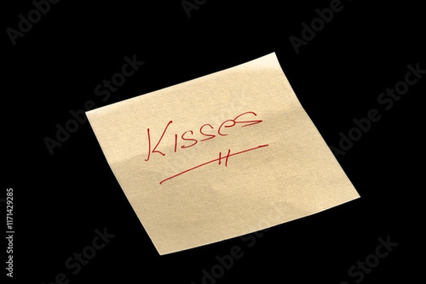 Fototapeta Yellow adhesive paper, similar to a Post-it note, on a black background with the words “Kisses” handwritten in red ink.