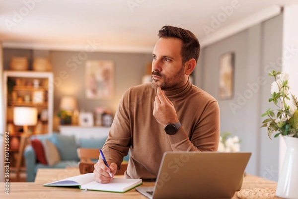 Fototapeta Portrait of serious male programmer thinking and writing in note pad while working over laptop at home