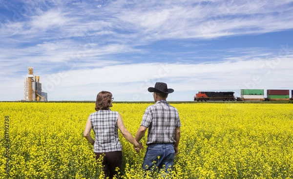Obraz horizontal image of a husband and wife checking canola field while a train rumbles by in the background next to a grain elevator.
