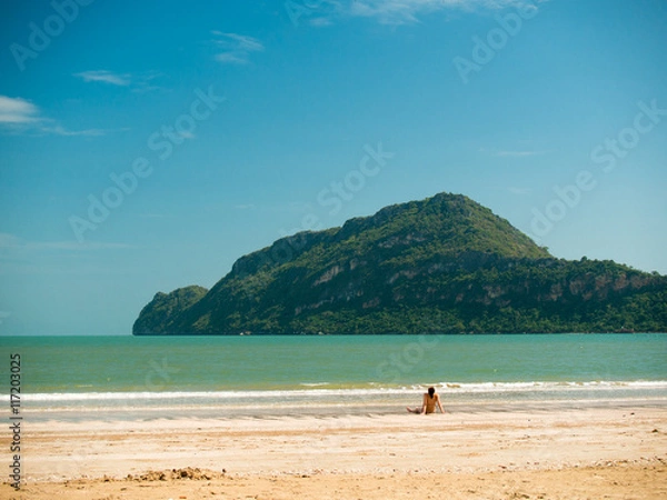 Fototapeta Toned image of a woman sits on the beach during low tide on the background of the islands with trees and sky with clouds