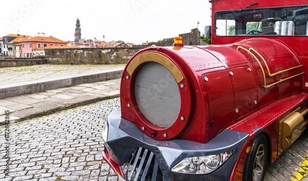 Fototapeta A vibrant red touristic train on the cobbled streets of Porto, with the iconic Clerigos Tower in the background under a soft overcast sky.