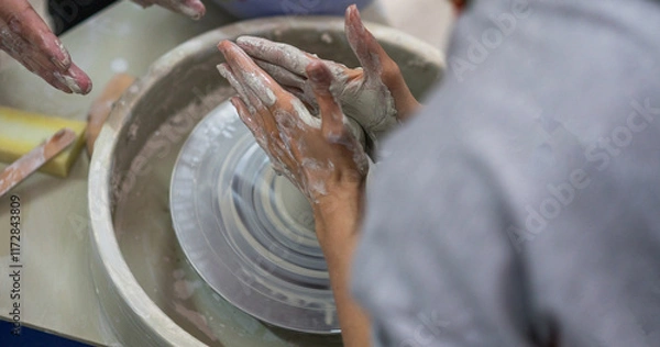 Fototapeta Close-up of a person's hands shaping clay on a pottery wheel. The individual's hands are coated in wet clay, meticulously molding the material. The process takes place in a pottery studio.