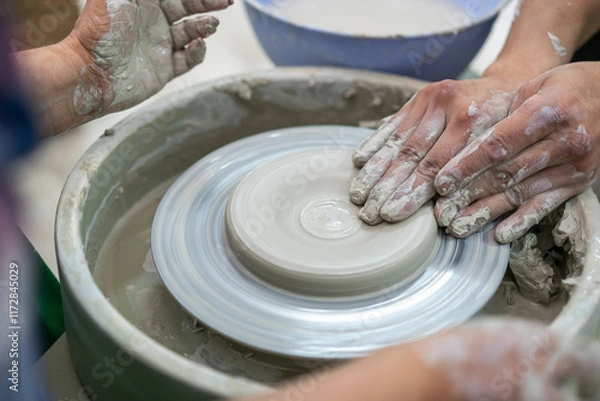 Fototapeta Close-up of hands covered in wet clay as two people collaboratively shape a pottery piece on a spinning wheel, emphasizing the teamwork and artistry involved in ceramic craftsmanship.