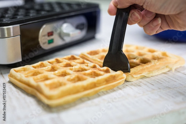 Obraz Mit einem schwarzen Spatel werden auf einer weissen Holzplatte zwei Waffeln geteilt. Im Hintergrund steht ein eckiges Waffeleisen.