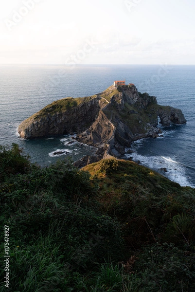 Fototapeta A church in the sea. Long winding steps leading to a church built on a cliff in the ocean. The sun is setting creating an orange glow over the scene, which contrasts with the blue ocean.