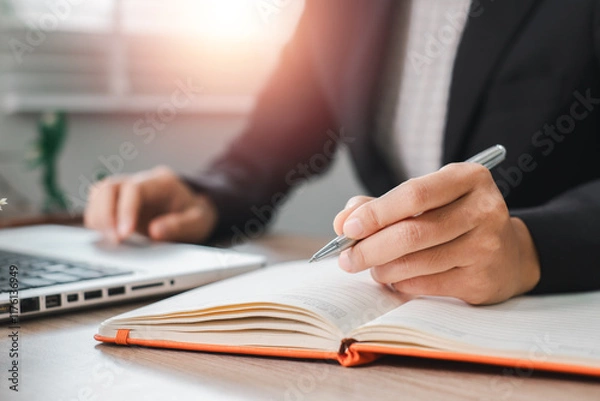 Fototapeta Close-up of a business professional writing in a notebook at a desk with a laptop, illuminated by soft sunlight.