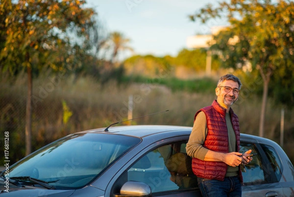 Fototapeta Smiling man leaning on a car during golden hour in an outdoor setting