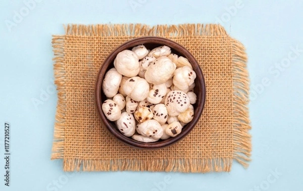 Obraz Top View of Fox Nut or Makhana in a Wooden Bowl on Burlap Fabric Isolated on Light Blue Background with Copy Space, Also Known as Lotus Seed Pop, Euryale Ferox or Gorgon Nut