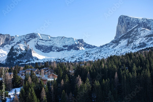 Fototapeta Vallée enneigée entre au pied d'une montagne avec village de station de ski niché au milieu d'une forêt de sapins.