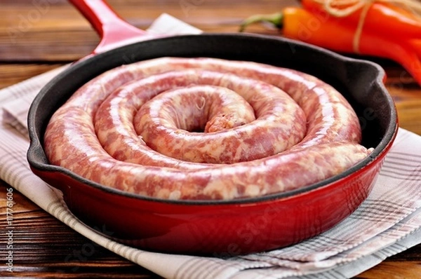 Fototapeta Raw pork sausage in a cast iron frying pan on a wooden background.