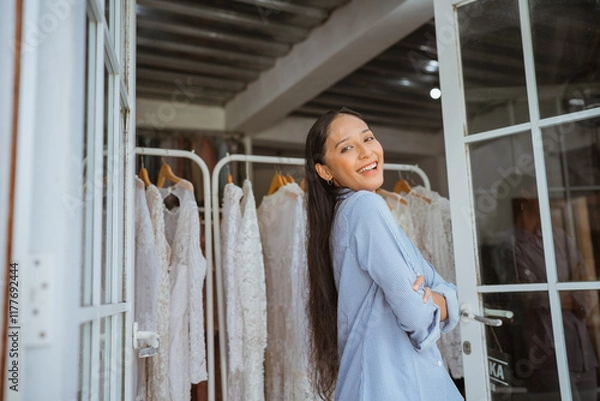Obraz A smiling woman dressed in a light blue attire poses gracefully in front of elegant gowns