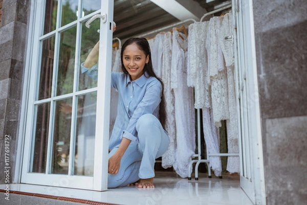 Obraz A smiling woman poses in a doorway with clothes hanging nearby, creating a joyful vibe