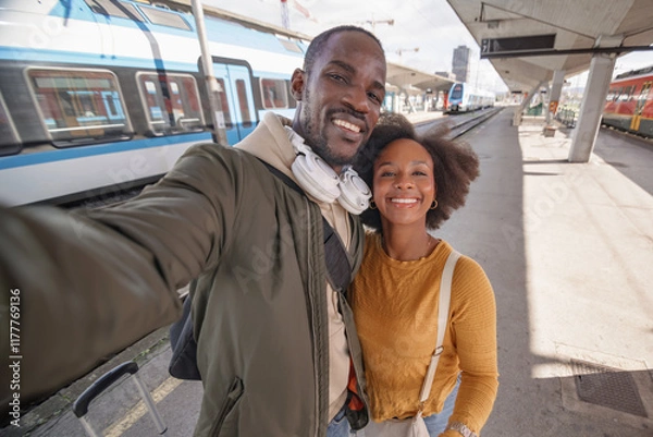 Fototapeta Joyful African American couple taking a selfie at a railway station before embarking on their train journey together. Travel happiness concept.