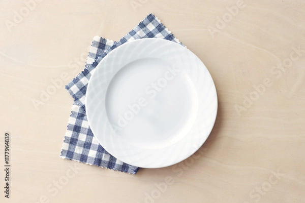 Fototapeta Empty white plate with blue fabric napkin on wooden table. Top view.	