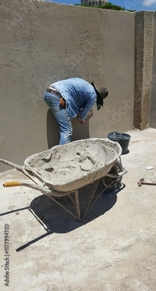 Fototapeta Pedreiro no trabalho de construção do muro de cimento, com carrinho de massa de cimento ao lado.