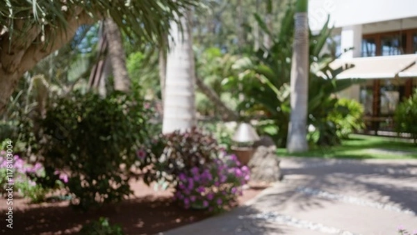Fototapeta Blurred background of a tropical resort garden with vibrant flowers, palm trees, and lush greenery.