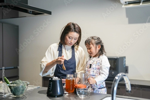 Fototapeta A Japanese parent and child, a woman in her 30s, a man in his 20s, and a 6-year-old girl, are making juice by putting cut carrots and honey in a blender in their home kitchen.