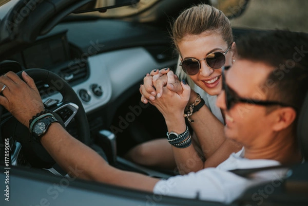 Obraz cheerful family couple lovers smiling and cheating to each other driving a cabriolet and searching the location they need by pointing on car gps system