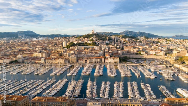 Fototapeta Aerial view of Marseille, France, showcasing the Old Port filled with yachts and the city's skyline, including Notre-Dame de la Garde on a hill under a partly cloudy blue sky