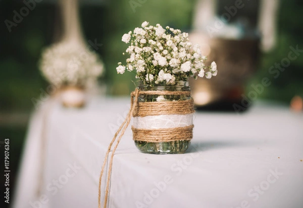 Fototapeta Wedding bouquet of white small flowers in a glass vase on a white fabric cotton tablecloth with green background.
