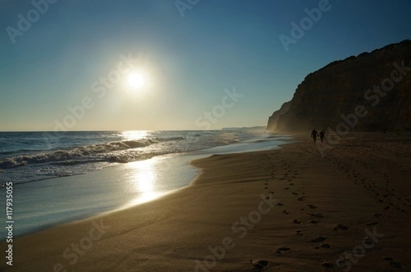 Fototapeta A couple strolls along the water's edge at high tide leaving two lines of foot prints leading down the beach. The sun is reflected on the water and on the wet sand. Algarve, Portugal.