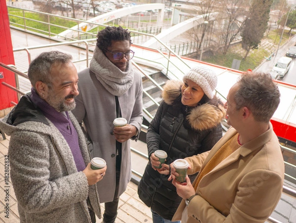 Fototapeta A group of four business colleagues taking a coffee break outdoors on a winter day. They are standing together, smiling, and holding coffee cups in an urban business district. Dressed in warm coats