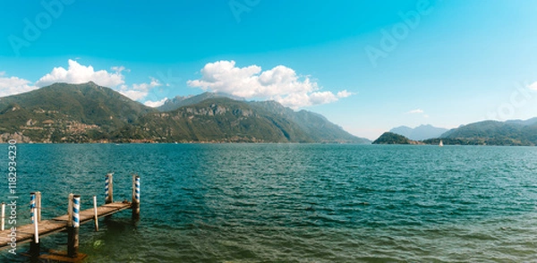 Obraz Tranquil scene of a wooden pier extending into the calm waters of Lake Como, surrounded by lush green mountains and a clear blue sky, creating a peaceful and scenic landscape in Menagg Lombardy, Italy