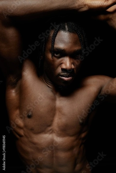 Fototapeta Portrait of tuned Black man with bare torso confidently posing against black background in dimly lit studio