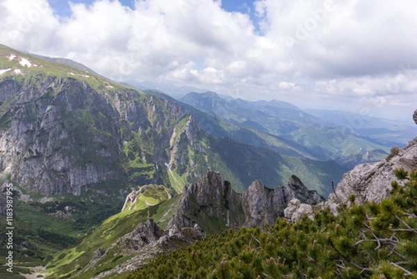 Fototapeta 
A mountain range with pointed peaks, steep slopes and deep valleys covered with sparse trees and shrubs, with lower mountain ranges visible in the distance.