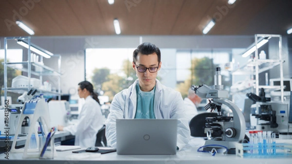 Fototapeta Genetic Research Scientist Working on a Laptop Computer in an Applied Science Laboratory. Portrait of a Handsome Young Multiethnic Lab Engineer in White Coat at Work
