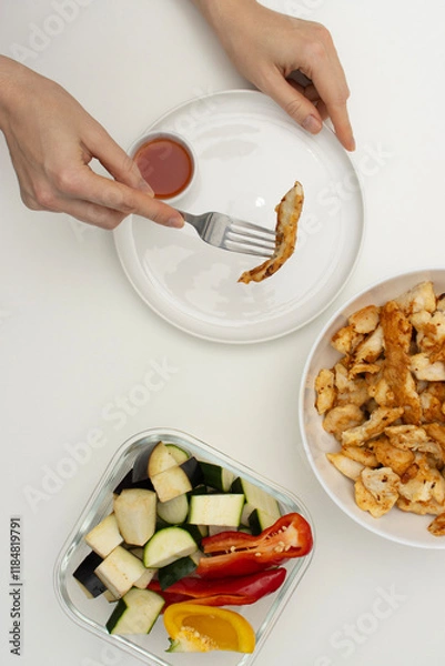 Fototapeta Female hand holding fork with piece of chicken. Homemade food on white table. Top view, copy space