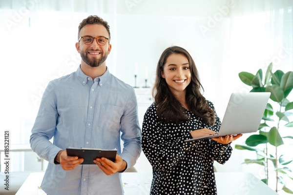 Fototapeta A man in a blue shirt holds a tablet while standing next to a brunette woman holding a laptop, confident colleagues looking at the camera and smiling standing in the modern office
