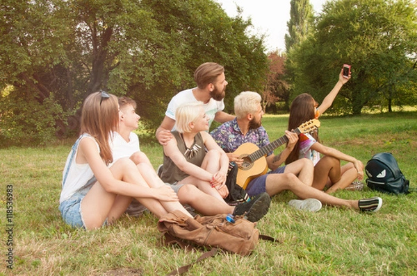 Fototapeta Group of smiling friends making selfie with smartphone camera in park