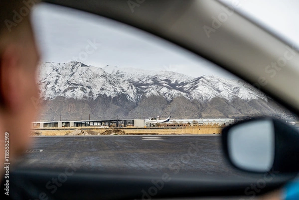 Fototapeta airplane taking off against a backdrop of snow-capped mountains, framed by the interior person driving car window