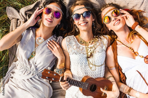 Fototapeta Trzy urocze hippie dziewczyny leżące na kratę na zewnątrz, najlepsi przyjaciele bawią się i śmieją, grają w ukulele, okulary przeciwsłoneczne, pióra we włosach, bransoletki, tatuaż flash, indie, Bohemia, widok z góry w stylu boho