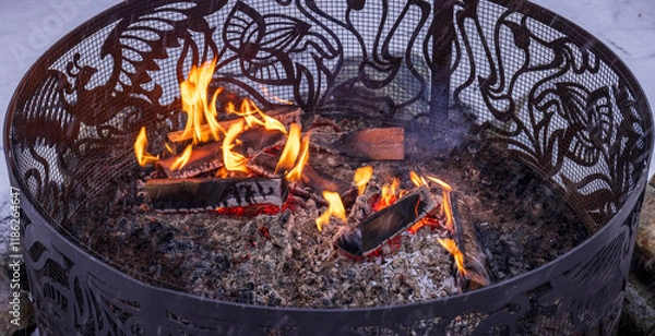 Obraz An intricately designed fire pit with decorative metalwork and burning flames, creating warmth and ambiance against a wintry backdrop of snow-covered ground and stones.