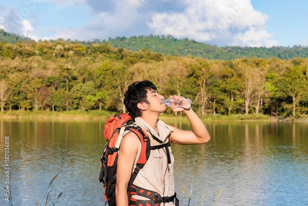 Fototapeta An asian man with backpacker smile and feeling fresh by drinking a bottle of water beside a lake in mountain alone
