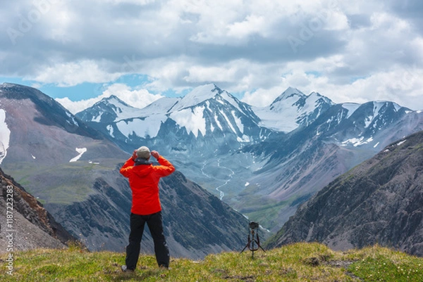 Fototapeta Man in vivid red jacket admire alpine scenery on sunlit green grassy hill near precipice edge. Guy on mountain pass enjoying view to few big snowy pointy peaks. Three large snow peaked tops far away.