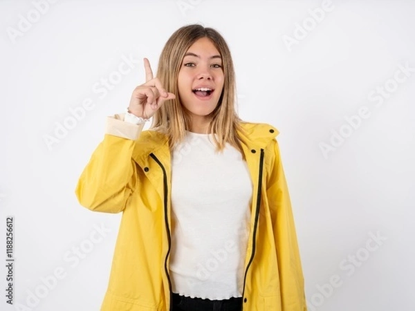 Fototapeta Beautiful teen girl wearing rain coat pointing finger up and looking inspired by genius thought, showing good idea sign, having clever solution in mind