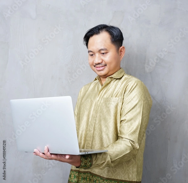 Obraz Adult Asian man wearing batik smiling at camera while using his laptop - Indonesia men